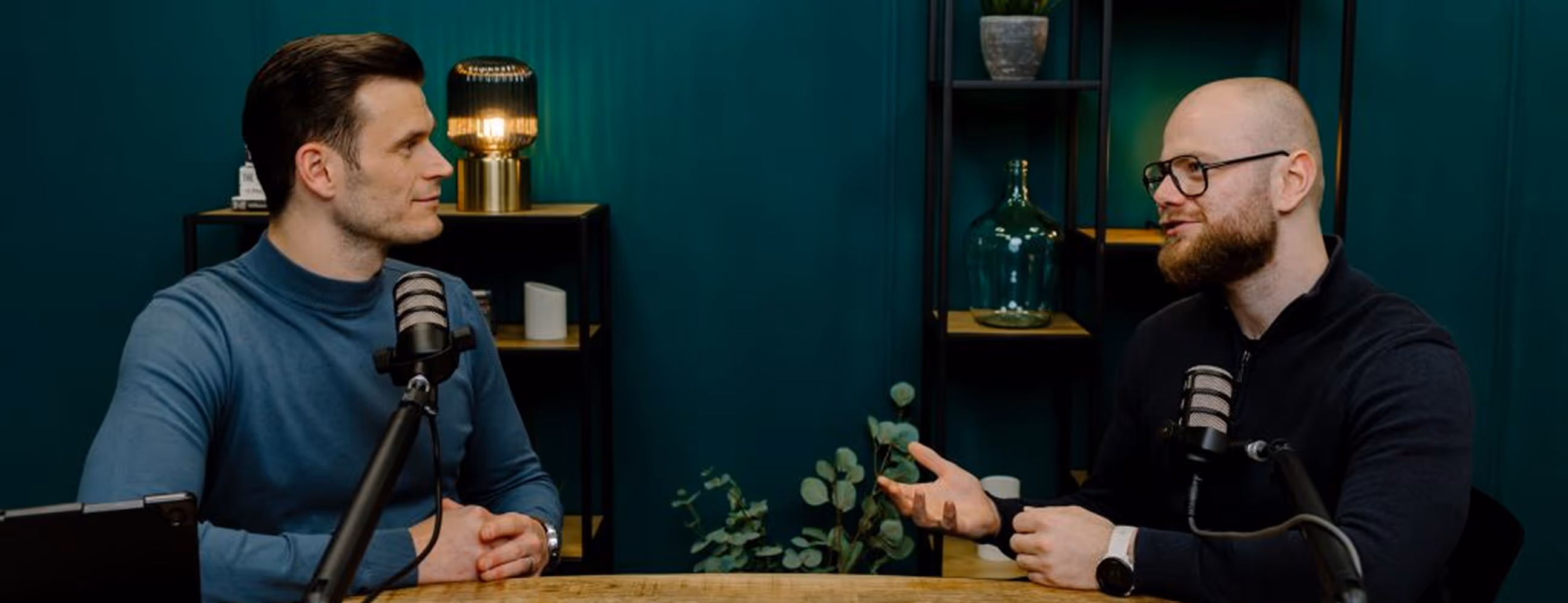 Twee mannen in gesprek aan tafel in de groene podcaststudio van Onruststokerz in Roermond, met microfoons op armen